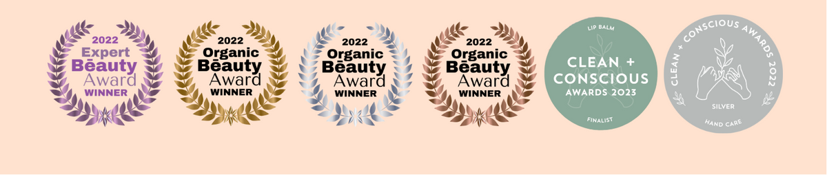 Six circular award seals for a beauty product arranged in a row on a pale peach background. The seals include 2022 Expert Beauty Award, 2022 Organic Beauty Award (twice, one gold, one blue), and Clean + Conscious Awards 2022.